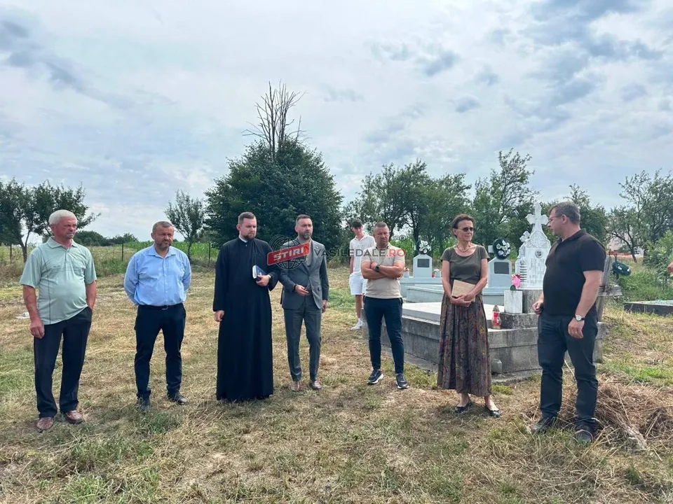 Un grup de șapte persoane stă în picioare pe un teren cu iarbă, lângă un monument de piatră sub un cer înnorat.