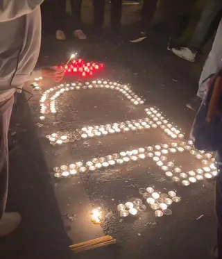 Multe lumânări aprinse sunt așezate pe asfalt în formă de litere, în timp ce o persoană ține o lumânare în mână.