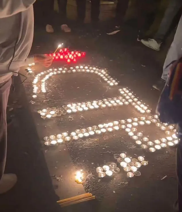 Multe lumânări aprinse sunt așezate pe asfalt în formă de litere, în timp ce o persoană ține o lumânare în mână.