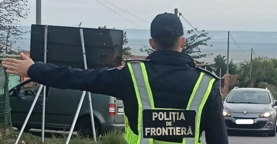 Un agent de poliție de frontieră cu vestă reflectorizantă stă cu spatele, întinzând brațul stâng spre un vehicul gri aflat pe drum.