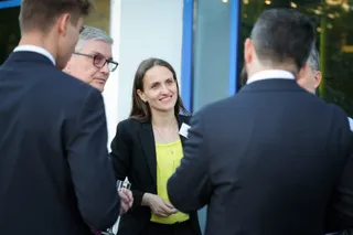 O femeie cu părul lung, purtând un sacou negru și un tricou galben, zâmbește în mijlocul unui grup de bărbați în costume închise la culoare.