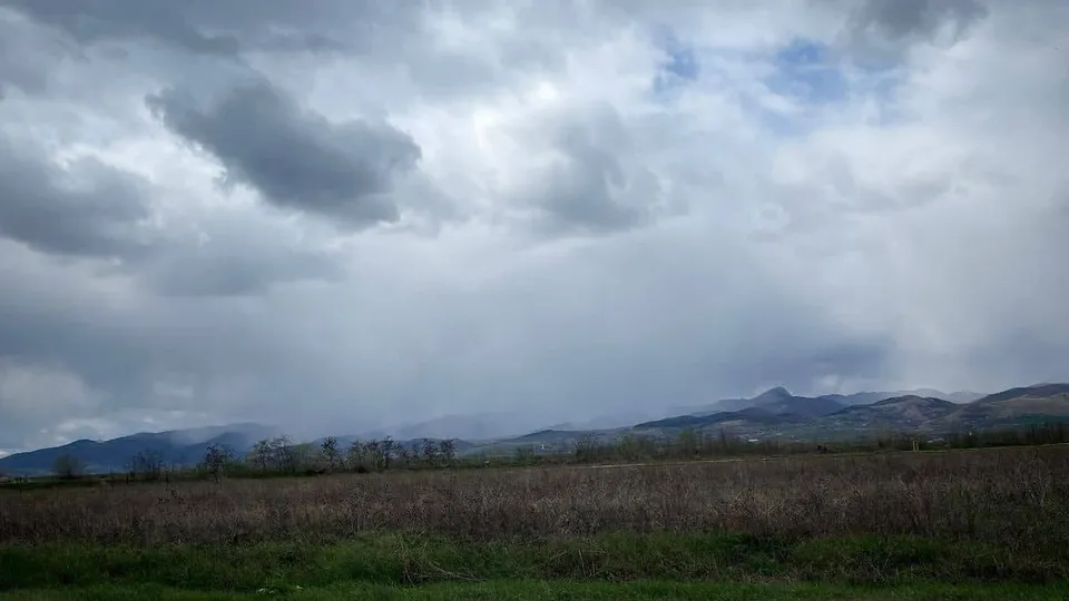 Un câmp cu vegetație uscată se întinde în prim-plan sub un cer acoperit de nori cenușii și zone de precipitații.