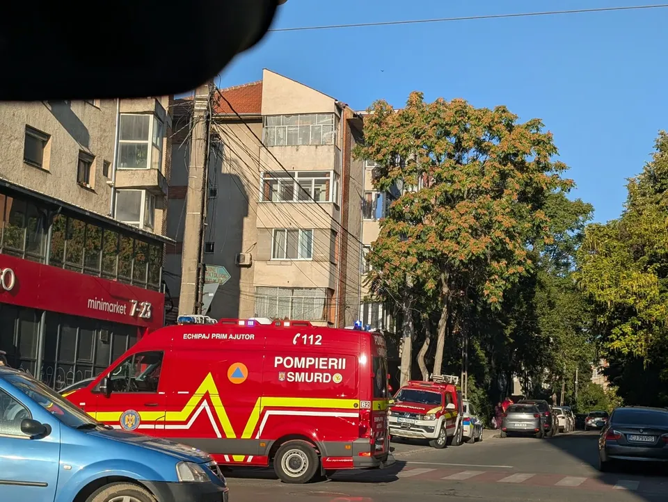 Un vehicul de serviciu roșu și galben cu inscripții de ajutor este parcat pe un drum lângă clădiri și copaci.