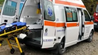 O ambulanță albă cu dungi portocalii este parcată cu portierele deschise, afișând un pat de transport medical pe exteriorul vehiculului.