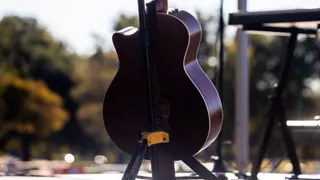 O chitară acustică este așezată pe un suport negru în fața unor copaci verzi și a unui suport de partitură într-un decor exterior.