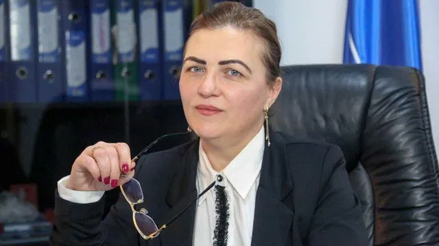 O femeie cu părul prins într-o coafură strânsă poartă un sacou negru și o cămașă albă, ținând ochelari cu ramă subțire în mână.