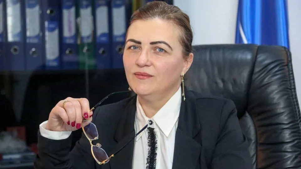 O femeie cu părul prins într-o coafură strânsă poartă un sacou negru și o cămașă albă, ținând ochelari cu ramă subțire în mână.