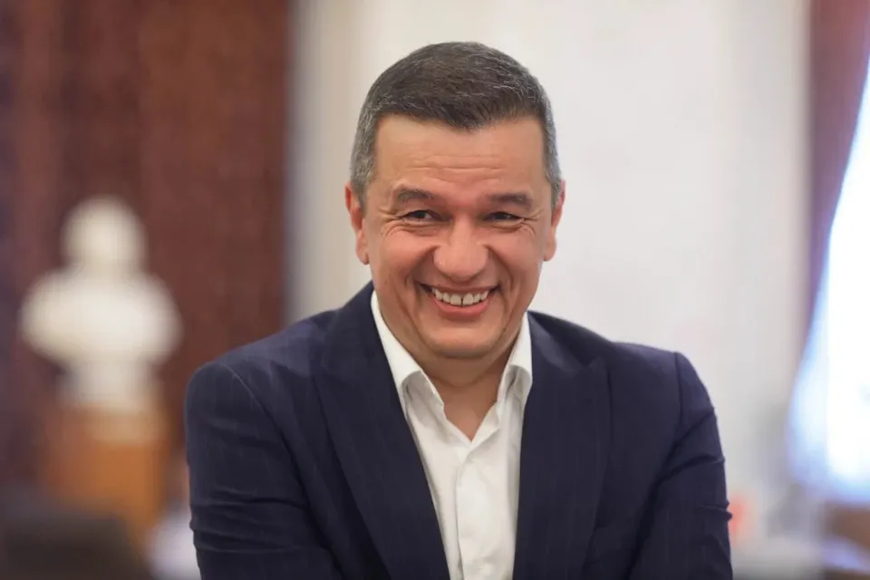 Un bărbat cu părul scurt și gri zâmbește, purtând o cămașă albă și un sacou închis la culoare.