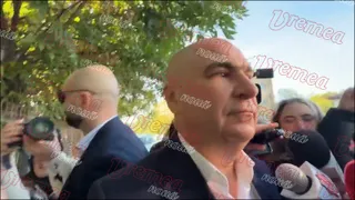 Un bărbat cu capul chel și cămașă albă este filmat de mai mulți oameni care țin microfoane și aparate foto în fața unui copac.