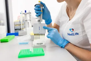 O persoană cu mănuși albastre manipulează un pipetaj într-un laborator, purtând o halat alb cu un logo pe buzunar.