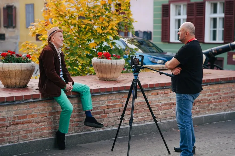 Un bărbat cu pălărie și sacou maro stă pe un zid de cărămidă, în timp ce un alt bărbat cu tricou negru filmează cu o cameră pe trepied.