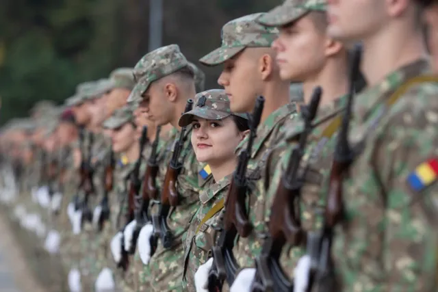 O linie de soldați în uniformă de camuflaj stau în rând, purtând căciuli militare și ținând puști cu accesorii albe.
