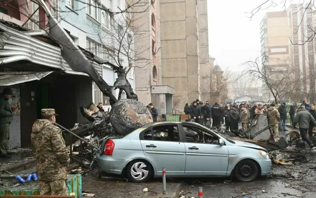 O mașină de culoare deschisă stă în fața unei structuri metalice distruse, lângă un grup de persoane și un individ în uniformă militară.