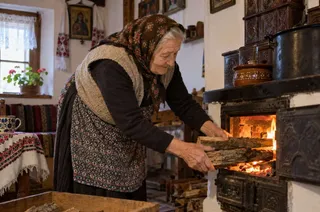 O femeie în vârstă cu batic în cap și vestă de lână pune lemne într-un cuptor de încălzit cu foc aprins.
