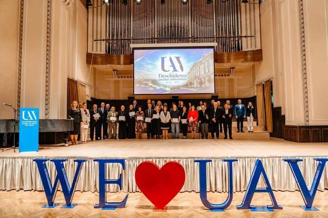 Un grup de persoane stă în rând pe o scenă de teatru în fața unui ecran mare care afișează un logo și un text.