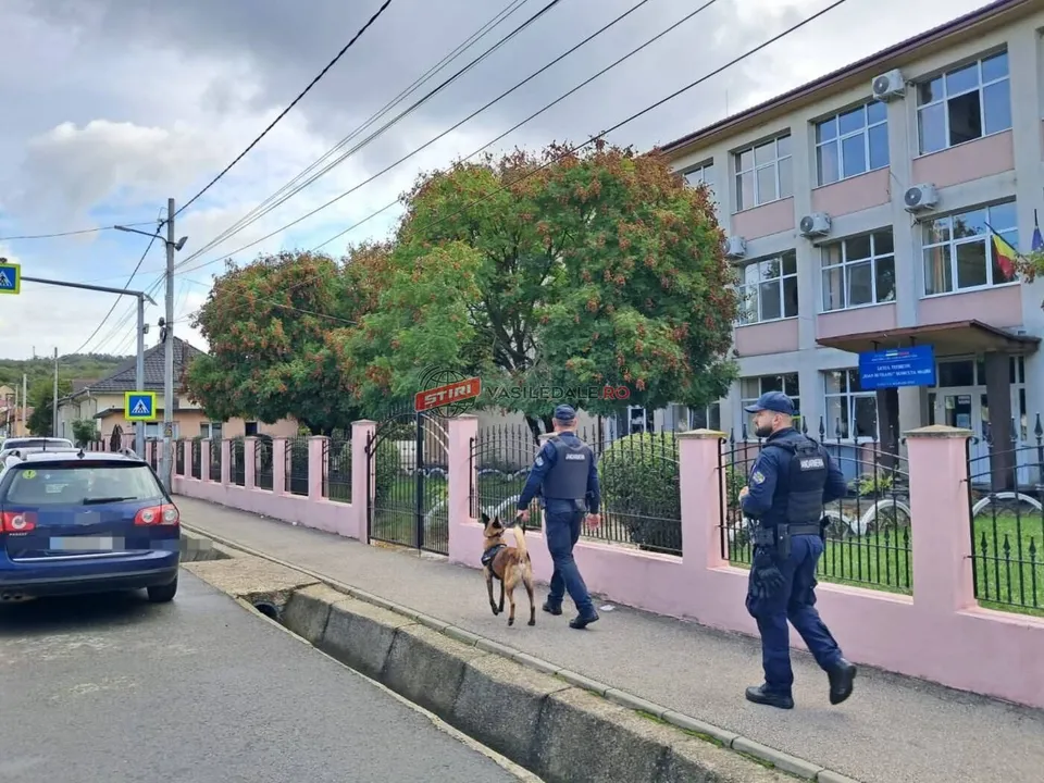 Doi polițiști în uniformă întunecată merg pe un trotuar lângă un gard roz, însoțiți de un câine de serviciu.