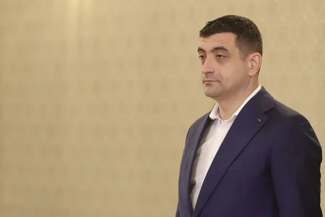 Un bărbat cu părul scurt și întunecat poartă un sacou albastru peste o cămașă albă, stând în fața unui perete de culoare deschisă.