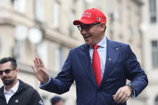 Un bărbat zâmbitor cu o pălărie roșie și ochelari negri poartă un costum albastru cu cravată roșie și salută cu mâna dreaptă.