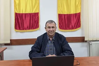 Un bărbat cu o cămașă în carouri și o jachetă închisă stă la o masă de lemn în fața a două steaguri galben și roșu.