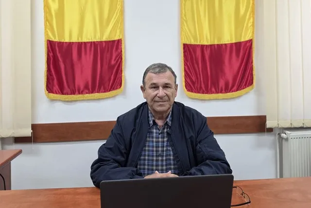 Un bărbat cu o cămașă în carouri și o jachetă închisă stă la o masă de lemn în fața a două steaguri galben și roșu.