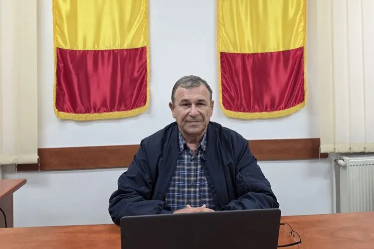 Un bărbat cu o cămașă în carouri și o jachetă închisă stă la o masă de lemn în fața a două steaguri galben și roșu.