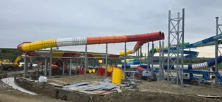Mai multe tobogane colorate de aquapark sunt instalate pe niște structuri metalice într-un șantier cu utilaje de construcții și pământ excavat.