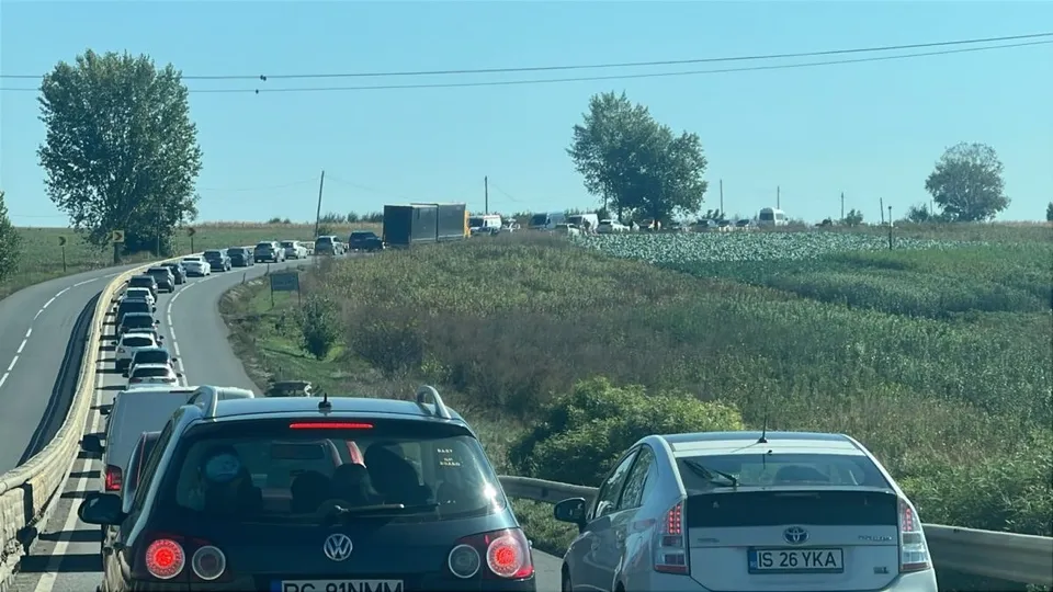 Două autoturisme circulă pe un drum asfaltat lângă un gard de protecție, în timp ce un rând de mașini este parcat pe marginea drumului.