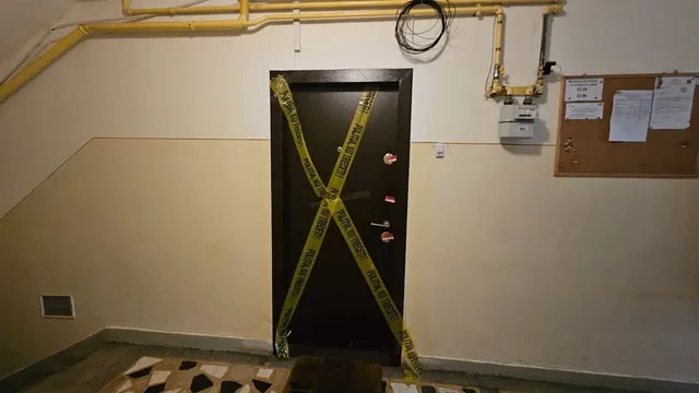 O ușă neagră este marcată cu benzi galbene în formă de X într-un interior cu pereți deschise și țevi galbene pe perete.