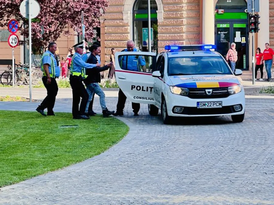 Patru bărbați stau lângă o mașină albă de poliție cu portierele deschise pe un trotuar lângă o zonă cu iarbă.