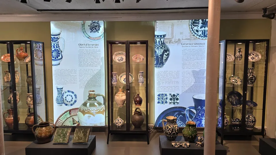 Diverse vase și obiecte din ceramică decorate sunt expuse în vitrine de sticlă și pe suporturi într-o încăpere de muzeu.
