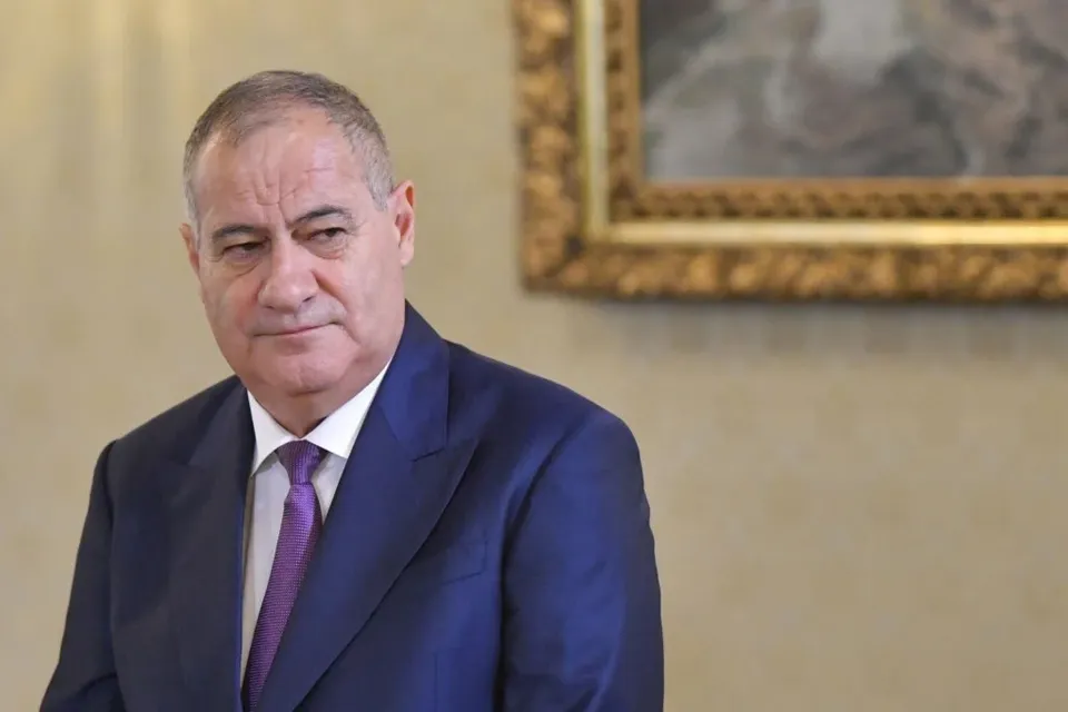 Un bărbat în costum albastru cu cravată mov este pozitionat în prim plan, cu un tablou în cadrul aurit în fundal.