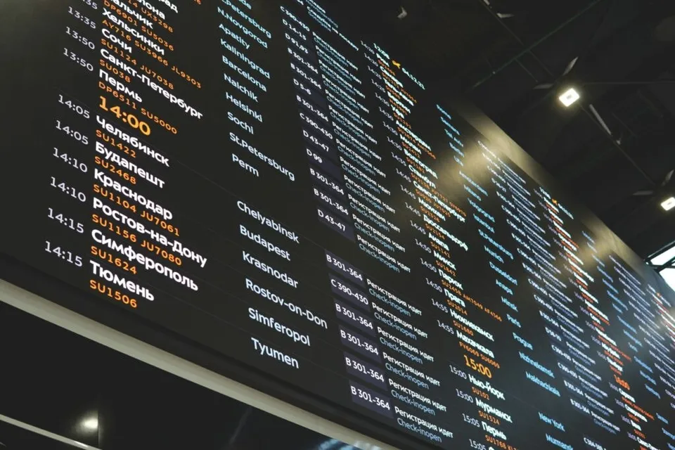 Un panou digital de informații afișează liste de destinații, ore și numere de zbor pe un fundal negru într-un spațiu interior iluminat.