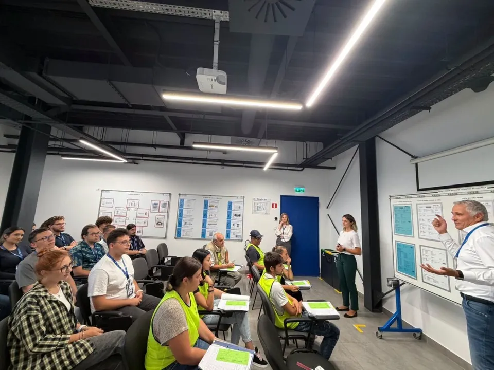 Un grup de persoane stă așezat la mese într-o sală de curs, privind spre un bărbat care vorbește în picioare în fața unui panou.