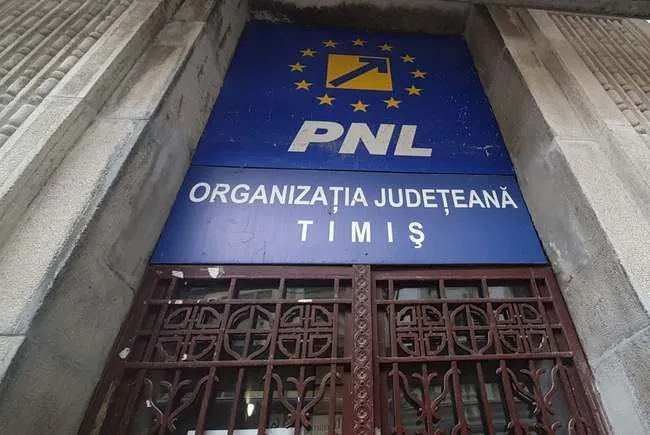 Un panou albastru cu textul alb și un logo galben este montat deasupra unei porți metalice maronii.
