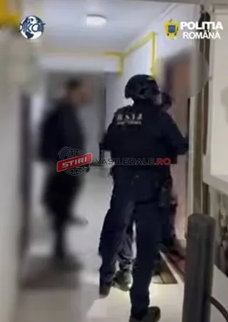 Un agent de poliție cu echipament tactic și cască neagră stă într-un hol lângă o ușă deschisă.