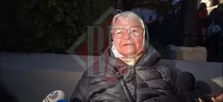 O femeie cu ochelari și o eșarfă pe cap poartă o geacă neagră și privește spre o cameră de filmare cu microfoane în prim-plan.