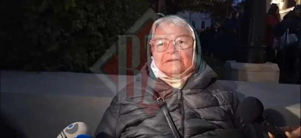 O femeie cu ochelari și o eșarfă pe cap poartă o geacă neagră și privește spre o cameră de filmare cu microfoane în prim-plan.