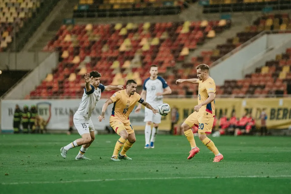 Patru jucători de fotbal sunt pe un teren verde, doi purtând echipamente galbene și doi echipamente albe, într-un stadion cu tribune colorate.
