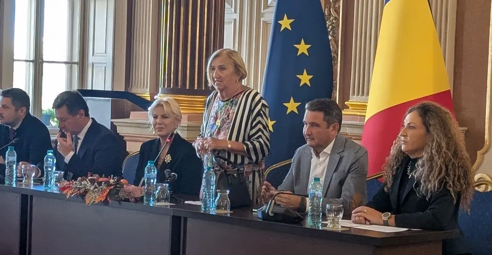 Șase persoane stau sau stau în picioare la o masă lungă cu sticle de apă și microfoane în fața unor steaguri.