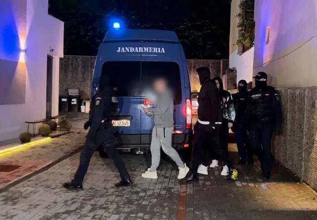 Patru persoane în uniformă închisă și un vehicul albastru cu inscripția Jandarmeria sunt poziționate pe un drum pavat lângă clădiri noaptea.