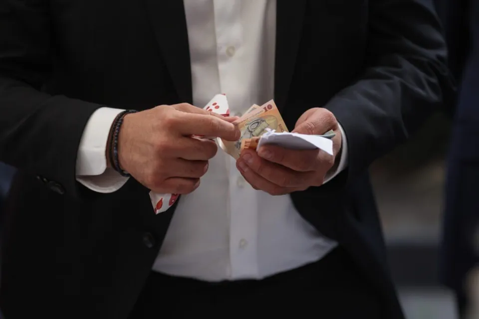 Un bărbat într-un costum negru și cămașă albă ține în mâini un teanc de bancnote colorate.
