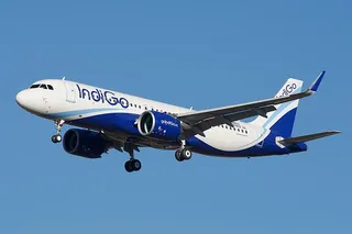 Un avion alb și albastru cu inscripția Indigo zboară pe un cer senin cu trenul de aterizare extins.