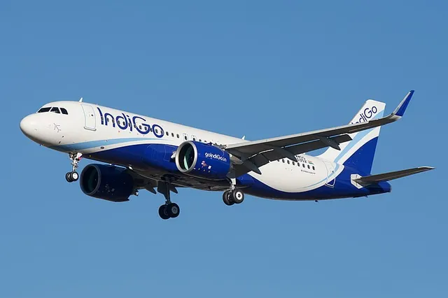 Un avion alb și albastru cu inscripția Indigo zboară pe un cer senin cu trenul de aterizare extins.