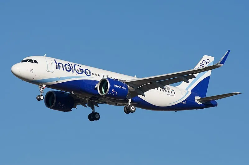 Un avion alb și albastru cu inscripția Indigo zboară pe un cer senin cu trenul de aterizare extins.