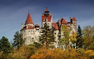 Un castel cu turnuri înalte și acoperișuri roșii este înconjurat de copaci cu frunziș verde și galben sub un cer albastru.