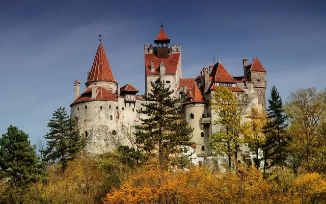 Un castel cu turnuri înalte și acoperișuri roșii este înconjurat de copaci cu frunziș verde și galben sub un cer albastru.