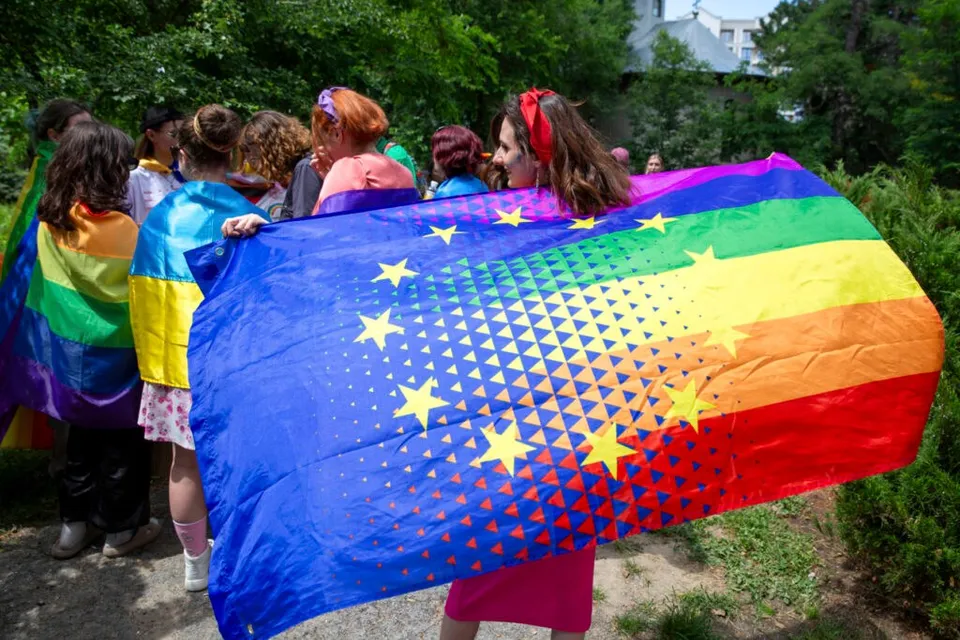 Un grup de persoane stă cu spatele și ține un steag mare care combină culorile curcubeului cu stelele galbenului pe un fundal albastru.