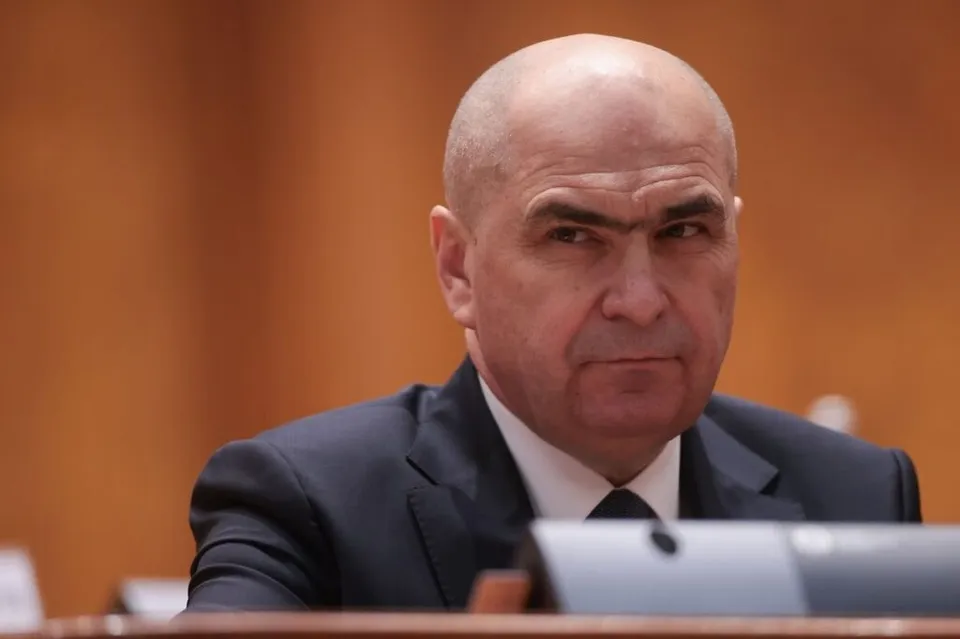 Un bărbat cu capul chel, purtând sacou închis la culoare și cămașă albă cu cravată neagră, privește spre lateral într-un decor cu fundal maro.