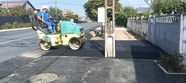 Un operator cu cască albă conduce un aparat de compactat pe o suprafață de asfalt proaspăt întins lângă un gard metalic.