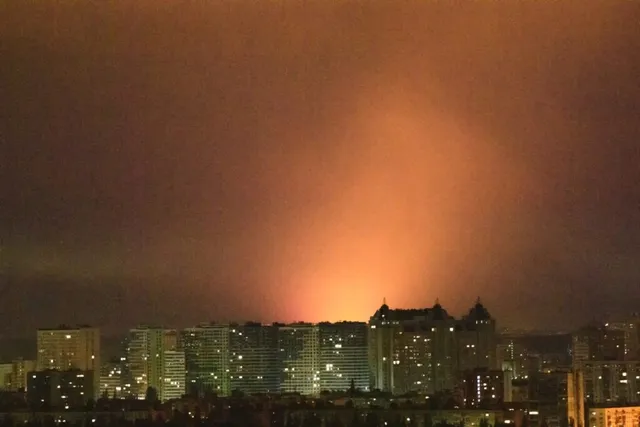 Lumina portocalie strălucește intens deasupra unei siluete de clădiri înalte sub un cer nocturn întunecat.